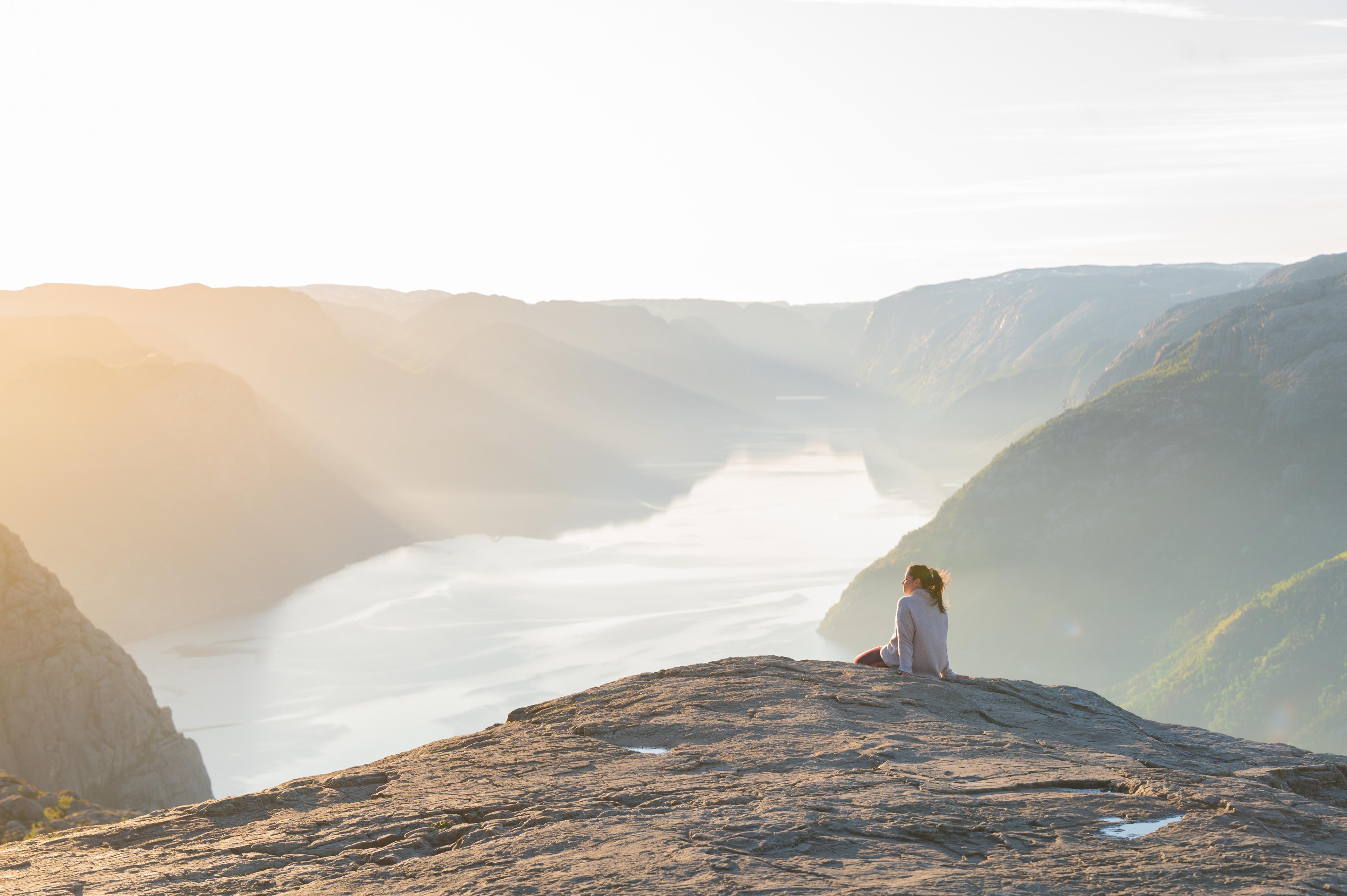 En dame sitter på kanten av Preikestolen-platået. HUn har utsikt over Lysefjorden!