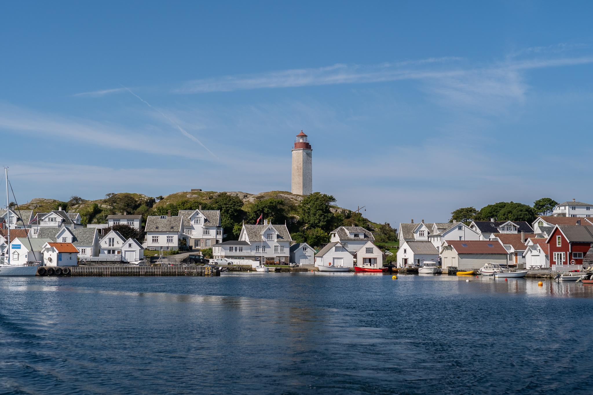 Øylandskap på Kvitsøy. Trehus og havneområdet på Ydstebø havn med Kvitsøy fyr i bakgrunnen. Sjøen i forgrunn.